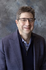 A studio portrait of Professor Eli Gabbay sitting in front of a mottled grey backdrop
