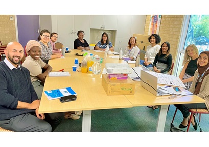A group of staff sit around a large board table in discussion. They are looking at the camera and smiling.
