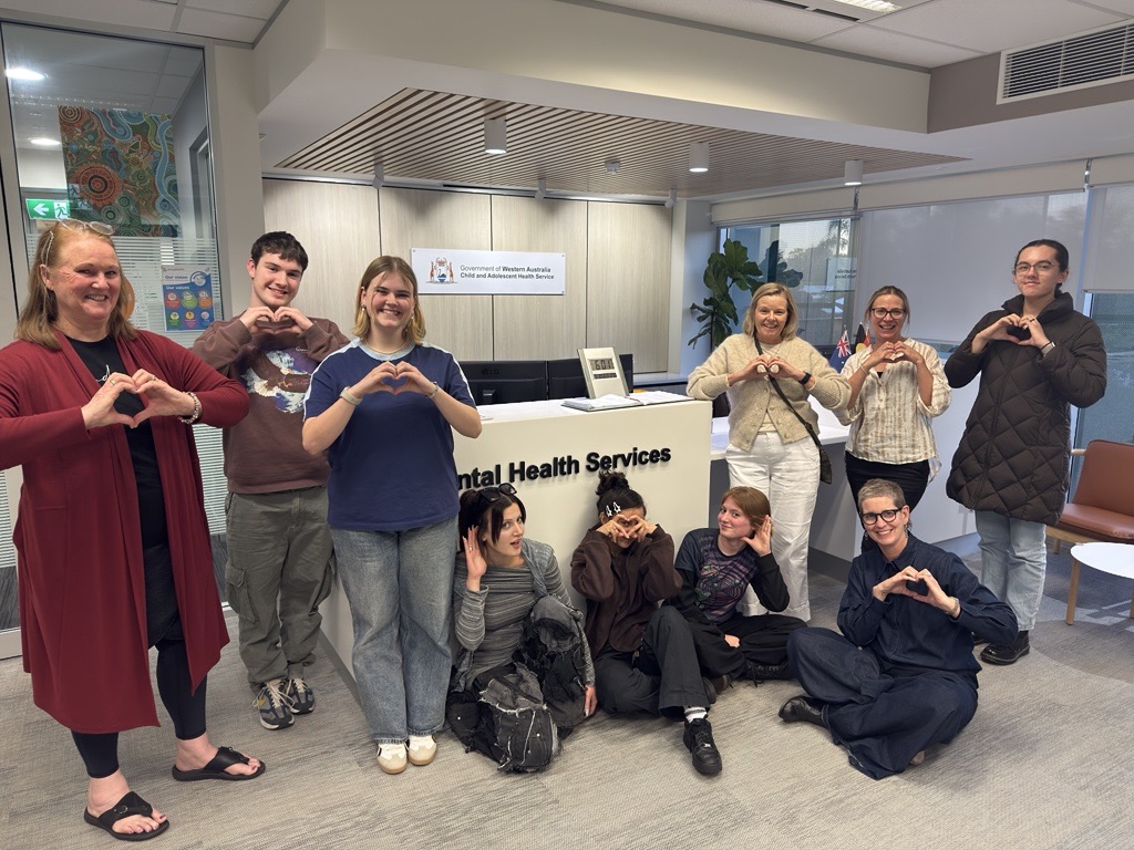 A group of ten people pose inside a modern building lobby near a reception desk. Several people are standing while others are seated or kneeling in front. Everyone is making a heart shape with their hands. Behind them is a light‑colored reception counter and a wall sign reading “Child and Adolescent Mental Health Services.” The space has polished flooring, overhead lighting, and a professional, welcoming interior.