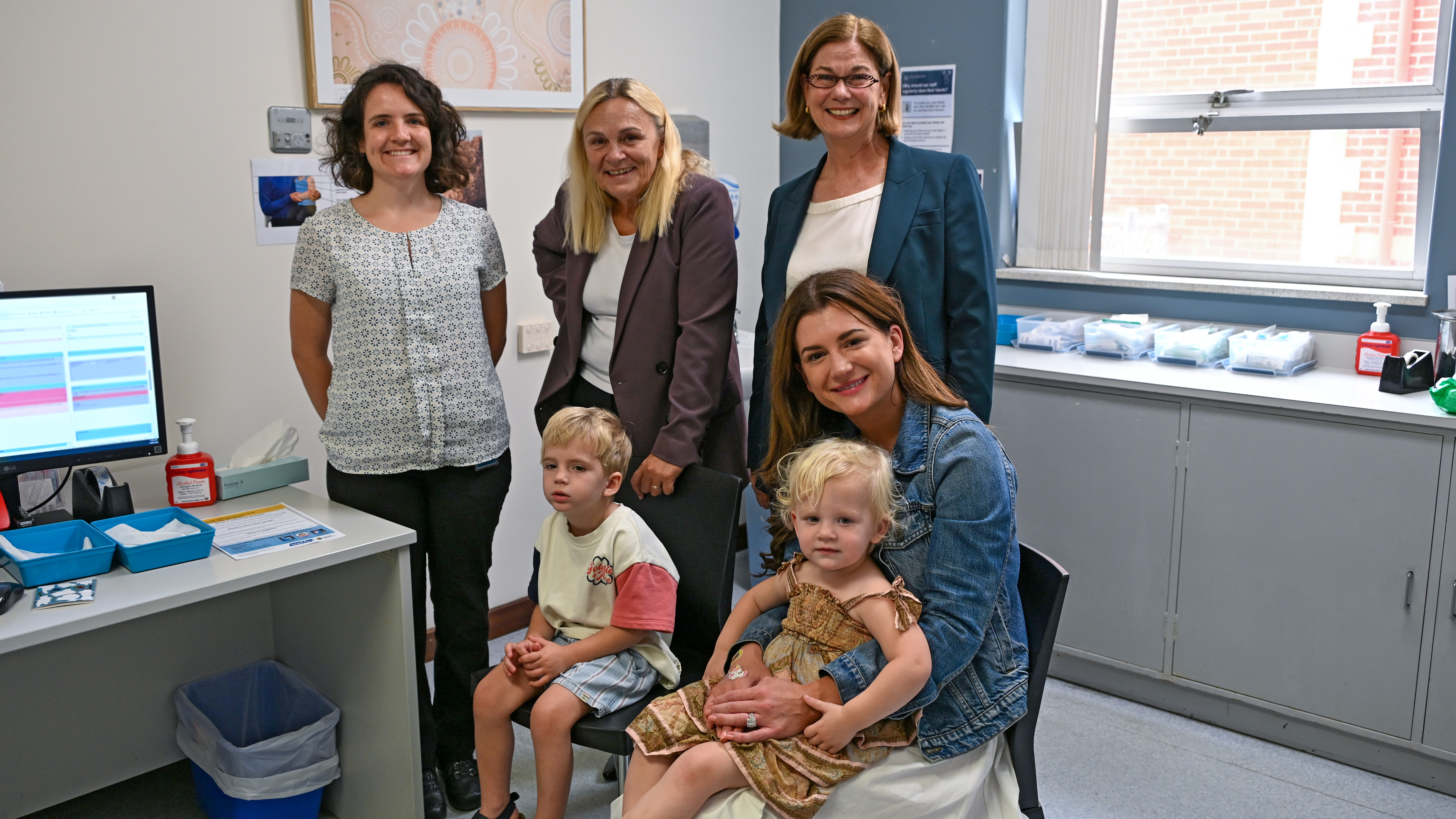A group of adults and two young children in a medical clinic room. The adults stand and sit around the children, who are seated on chairs. Medical equipment, supplies, a computer desk, cabinets, and a window are visible in the background.