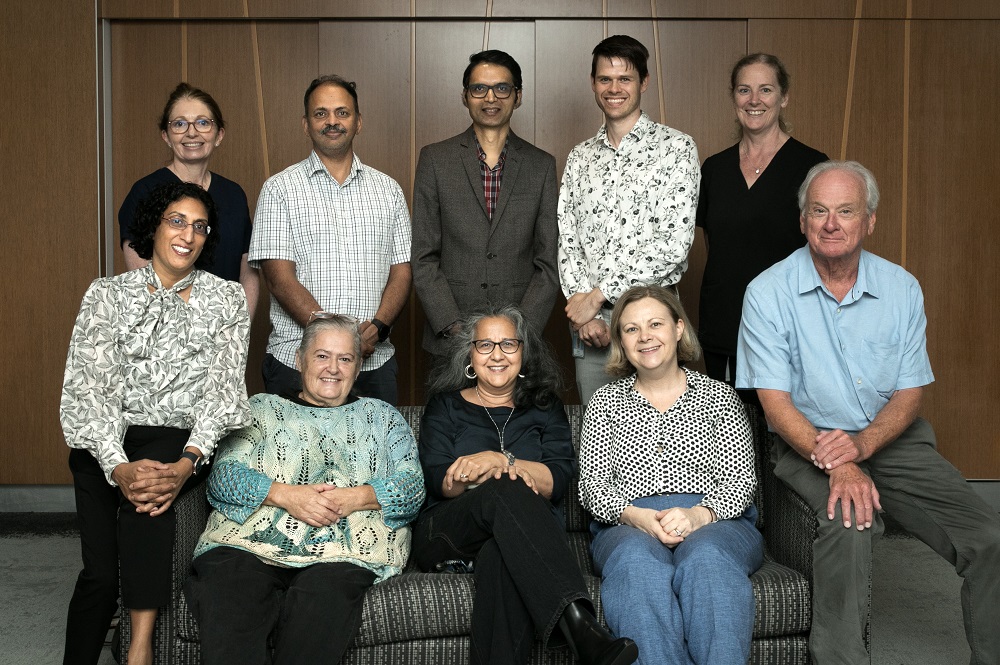 A group of tenadults posing for a formal group photo indoors. Five people are seated on a couch in the front row and five are standing behind them, all smiling toward the camera. They are dressed in business‑casual clothing, with a wood‑paneled wall in the background.