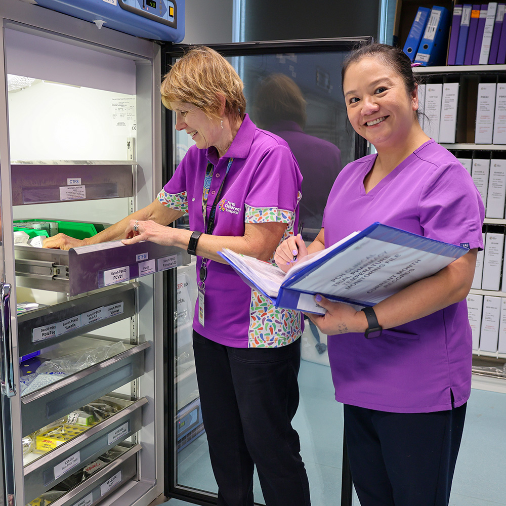 Two healthcare workers in purple uniforms stand beside an open medical refrigerator, with one person selecting labelled items from storage trays while the other holds a clipboard in a clinical storage room.